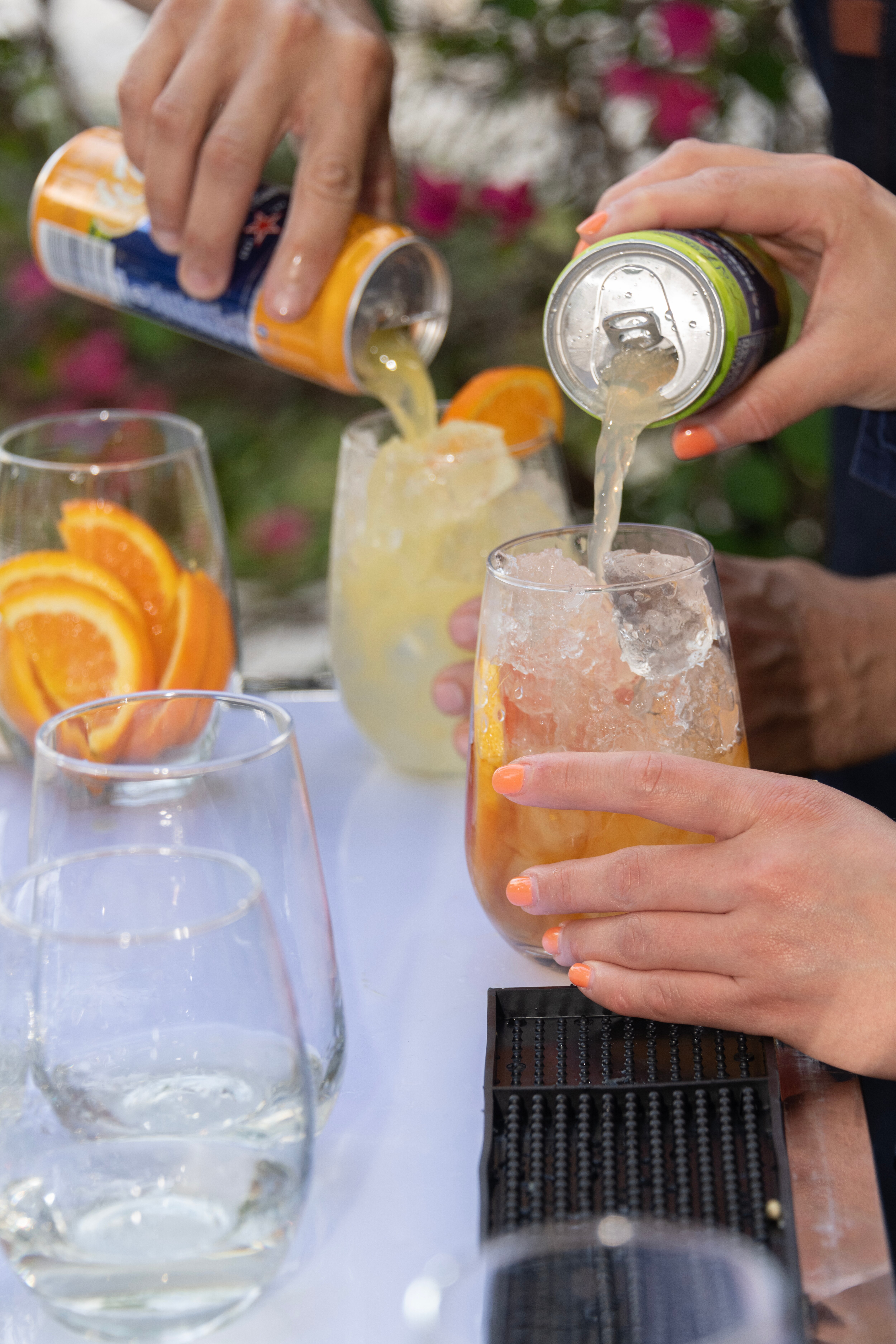 Hands pouring drinks from cans into glasses filled with ice and orange slices.